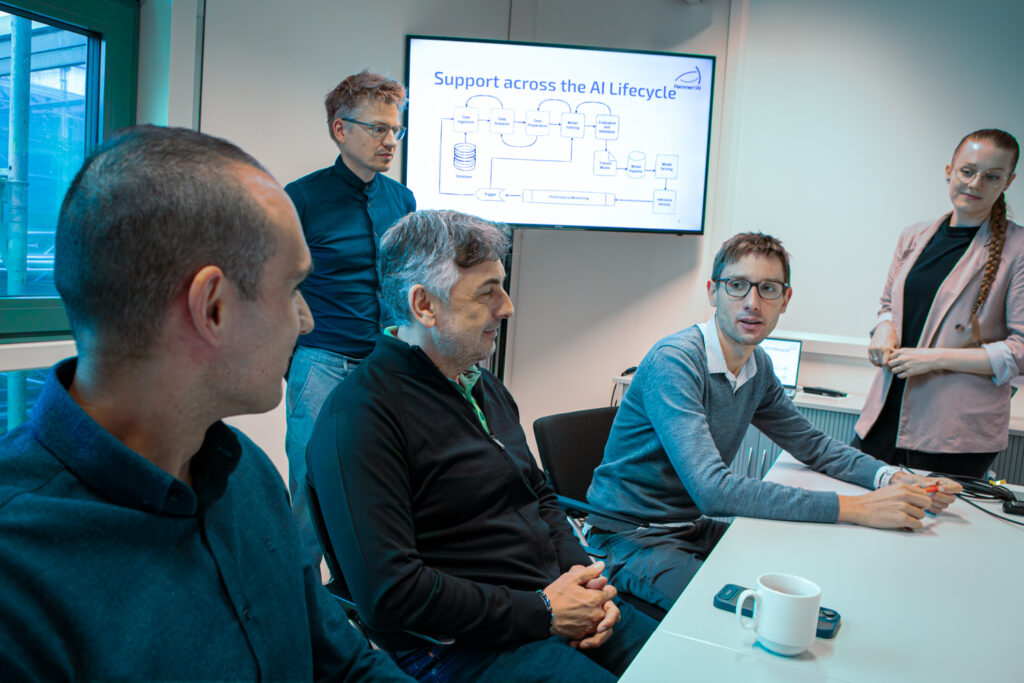 Fünf Personen diskutieren an einem Schreibtisch über etwas, während im Hintergrund auf einem Fernseher eine Präsentation zum KI-Lebenszyklus läuft.